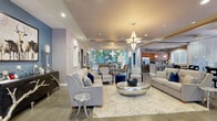 Located at 8200 Jayme Dr in Winter Garden, Florida, this single-story clubhouse offers a generously proportioned, thoughtfully designed gathering space spanning over 3,000 square feet. The interior is characterized by large-format tile flooring throughout, tray ceilings with recessed lighting, and an open, flowing layout that connects multiple functional zones seamlessly.

The main lounge area serves as the centerpiece of the space, featuring multiple seating arrangements accented by bold blue and neutral tones, statement geometric pendant lighting, and floor-to-ceiling glass walls that flood the interior with natural light and provide lush green views of the surrounding landscape. A secondary seating area with coordinated furnishings offers an inviting spot for casual gatherings.

The well-appointed kitchen is outfitted with warm wood cabinetry, granite countertops, stainless steel appliances including a full-size refrigerator and dishwasher, and a central island with bar seating — ideal for entertaining or community events. Adjacent to the kitchen, a dining area with high-top tables and crystal chandelier fixtures provides a stylish setting for meals.

A separate office or workroom is accessible from the main lounge, and large glass doors open directly to a covered outdoor patio overlooking a pool, seamlessly blending indoor and outdoor living.