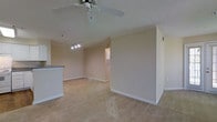 This single-floor residence offers a clean, functional layout designed for comfortable everyday living. The main living area is finished with wall-to-wall carpeting in a warm, neutral tone, creating a soft and inviting atmosphere. Crown molding runs along the upper perimeter of the walls, adding a classic architectural detail that elevates the overall finish of the space. The ceiling features a textured surface and is equipped with a ceiling fan, providing both air circulation and a practical lighting fixture. A track lighting fixture is also present, offering additional focused illumination to the living area. The walls are painted in a light, warm beige tone that reflects natural light and contributes to the open, airy feel of the room. A partial view of an adjacent counter — likely a kitchen peninsula or breakfast bar — is visible along one edge, suggesting a connected or open-concept kitchen layout nearby. A windowed door or pass-through is visible in the background, hinting at additional rooms or a private entry beyond. White baseboards provide a crisp contrast against the carpeted floors and neutral walls, maintaining a tidy and cohesive interior aesthetic throughout the space.