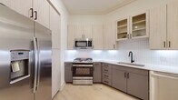 This contemporary single-floor residence showcases a cohesive modern design with light hardwood flooring and a neutral palette carried throughout. The kitchen is a standout space, featuring a two-tone cabinet scheme pairing light wood upper cabinetry with warm gray lower cabinets, all accented by matte black hardware. A crisp white subway tile backsplash, stainless steel appliances including a gas range with overhead microwave, full-size refrigerator, and an undermount sink with a sleek black faucet complete the well-appointed culinary space. Glass-front upper cabinets add an open, airy quality to the room. The bedroom offers a calm and restful retreat with soft neutral walls, natural light filtering through slatted blinds, and a connecting walk-in closet for generous storage. The bathroom is finished with large-format light tile flooring, a gray vanity with a white countertop, a tall matching storage tower, a framed mirror, and matte black fixtures throughout. A versatile living area benefits from elevated ceilings, recessed lighting, a ceiling fan, and French doors that open to the outdoors, inviting natural light and seamless indoor-outdoor flow. Together, these spaces form a refined and functional modern home.