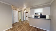 This single-story residence offers a practical and well-appointed layout with cohesive finishes throughout. Wood-look plank flooring runs through the main living areas, complemented by crown molding and textured ceilings that add a classic touch. The open living room is a standout, featuring a white brick corner fireplace, track lighting, and a large sliding glass door fitted with vertical blinds that allows natural light to fill the space. A ceiling fan overhead enhances comfort in the main area. The kitchen is positioned adjacent to the living space in a peninsula-style layout, offering dark granite-look countertops, white cabinetry, and black appliances including an electric range and refrigerator. A built-in pantry cabinet and under-cabinet ventilation hood add both functionality and storage. A stacked washer/dryer unit is conveniently tucked into a dedicated nook along the hallway. The bedroom features plush carpeting, crown molding, a ceiling fan, and a window with blinds providing natural light. The bathroom is outfitted with a white vanity, dark speckled countertop, dual mirrors, and white cabinetry. Throughout, the neutral gray color palette and consistent finishes create a clean, unified aesthetic.