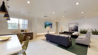 This thoughtfully designed single-floor amenity space offers a refined blend of comfort and functionality across several distinct areas. The main lounge serves as the centerpiece, featuring a generous seating arrangement with a patterned sofa, high-back accent chairs, and a tufted leather ottoman. Recessed lighting illuminates the crisp white ceilings, while curated artwork — including a striking geometric mountain landscape — adds character to the walls. Subtle green carpet accents anchor the seating area, complemented by elegant table lamps and decorative accessories that lend a warm, sophisticated ambiance. A wall-mounted flat-screen television and a built-in illuminated stone accent wall enhance the media area, making it ideal for both relaxation and social gatherings. A wet bar with dark wood cabinetry provides convenient service for entertaining. The full kitchen is exceptionally well-equipped, showcasing dark wood cabinetry, white quartz countertops, a mosaic tile backsplash, stainless steel appliances including a range with a wall-mounted hood, and a central island with seating. Abundant natural light pours in through large windows. Completing the space is a tranquil reading nook featuring three recessed diamond-pattern bookshelves with warm interior lighting, paired with plush high-back chairs and matching ottomans — a perfect retreat for quiet reflection.