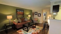 This single-floor residence offers a comfortable and stylishly curated living environment with a cohesive design aesthetic throughout. The living room serves as the centerpiece of the space, anchored by a rich, earth-toned sectional sofa adorned with patterned throw pillows that complement the warm color palette. A bold olive green accent wall adds depth and character to the room, while a large framed landscape painting serves as a striking focal point. A glass-topped coffee table sits atop a vibrant floral area rug, introducing color and texture to the hardwood flooring beneath. Soft lighting from a classic table lamp creates an inviting ambiance, and decorative branches in a tall vase add a natural, organic touch. A patterned accent chair with coral cushions provides additional seating, and wall-mounted artwork continues the decorative theme. Bar-height stools near the edge of the room suggest a casual dining or counter area nearby. A hallway visible from the living area leads to additional rooms, hinting at a well-connected and functional floor plan. Crown molding details along the ceiling add a refined architectural finish to the overall space.