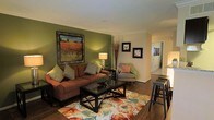 This single-floor residence offers a comfortable and stylishly curated living environment with a cohesive design aesthetic throughout. The living room serves as the centerpiece of the space, anchored by a rich, earth-toned sectional sofa adorned with patterned throw pillows that complement the warm color palette. A bold olive green accent wall adds depth and character to the room, while a large framed landscape painting serves as a striking focal point. A glass-topped coffee table sits atop a vibrant floral area rug, introducing color and texture to the hardwood flooring beneath. Soft lighting from a classic table lamp creates an inviting ambiance, and decorative branches in a tall vase add a natural, organic touch. A patterned accent chair with coral cushions provides additional seating, and wall-mounted artwork continues the decorative theme. Bar-height stools near the edge of the room suggest a casual dining or counter area nearby. A hallway visible from the living area leads to additional rooms, hinting at a well-connected and functional floor plan. Crown molding details along the ceiling add a refined architectural finish to the overall space.