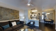 This single-floor residence offers a seamless open-concept layout that connects the living area and kitchen into one cohesive, inviting space. The living room is anchored by a dark, plush sofa accented with decorative pillows and paired with a sleek armchair, all arranged around a glass coffee table atop a textured area rug. A large abstract artwork adds a bold focal point to the wall, complementing the modern aesthetic. A ceiling fan with an integrated light fixture sits overhead, enhancing both comfort and style. The kitchen is defined by a raised breakfast bar that serves as a natural divider between the two spaces, topped with a light countertop and equipped with stainless steel appliances, including a full-sized refrigerator. Pendant lighting in contrasting black tones hangs above the kitchen area, adding a striking design element alongside under-cabinet illumination. The dark hardwood flooring runs throughout, grounding the space with a rich, contemporary feel. Neutral wall tones keep the interior feeling bright and open, while natural light filters in from what appears to be a windowed area, contributing to the overall airy ambiance of this well-appointed residence.