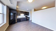 This single-floor space showcases a thoughtfully designed open-concept layout that seamlessly connects the kitchen and living area. The kitchen is outfitted with dark espresso cabinetry that extends from base to upper units, providing generous storage throughout. A full suite of black appliances — including a refrigerator, electric range, built-in microwave, and dishwasher — complements the cabinetry for a cohesive, modern aesthetic. Light-toned countertops offer a subtle contrast against the dark cabinetry, while under-cabinet lighting adds functional illumination to the workspace. A stainless steel sink is positioned along the countertop run, completing the kitchen's practical layout. Dark hardwood-style flooring flows continuously throughout the space, anchoring the design with a warm yet contemporary feel. The adjoining living area is open and generously proportioned, with clean white walls and recessed lighting overhead. Flush-mount ceiling fixtures provide additional ambient light. Crisp white baseboards trim the perimeter, adding a clean finishing detail. A hallway visible from the main space suggests additional rooms beyond, rounding out the functional layout of this well-appointed unit.