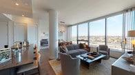 This single-floor high-rise residence showcases a modern design aesthetic defined by clean lines, quality finishes, and an abundance of natural light. The open-concept living area is anchored by floor-to-ceiling windows that frame sweeping panoramic city views, filling the space with daylight. A structural column adds architectural character, while a contemporary chandelier and warm wood flooring contribute to the refined atmosphere.

The living room accommodates generous seating arrangements centered around a low-profile coffee table, with ample room for both relaxing and entertaining. Adjacent to the living area, the dining space features a substantial dark-wood table paired with tan leather chairs beneath a sculptural multi-globe pendant light.

The kitchen is outfitted with dark cabinetry, a large center island with dark stone countertops, stainless steel appliances including a gas range and double ovens, and light hardwood flooring—offering a striking contrast of tones.

The bedrooms are serene and well-proportioned, with carpet flooring, curated built-in shelving, and large windows overlooking the cityscape. The bathrooms feature dramatic dark marble-style tile flooring, sleek vanities, and minimalist fixtures, completing the residence's cohesive contemporary character.