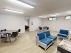 Located at 2067–2077 Prospect St in Burlington, Ontario, this shared laundry facility offers a well-organized, functional space spread across a single floor. The laundry area is equipped with a row of modern front-loading washers and dryers finished in white, set against clean white-tiled walls accented with a subtle light-blue tile border. A central folding counter with seating provides a convenient workspace for residents, while instructional signage is posted throughout for ease of use. The flooring throughout is a warm-toned light wood laminate that adds a clean, welcoming feel to the utilitarian space. Adjacent to the laundry area, a generously sized lounge offers a comfortable place to wait, furnished with contemporary blue-and-white seating including a sofa, armchairs, and a coffee table, along with a round dining table and chairs. The lounge is lit by overhead fluorescent track lighting and benefits from natural light filtering in through a row of high transom windows along one wall. The space is practical and well-maintained, with a clearly marked exit and open layout that encourages ease of movement throughout.