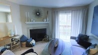 This single-floor living space offers a welcoming and well-appointed interior anchored by a classic tile-surround fireplace with a crisp white mantel, which serves as the room's focal point. The mantel is styled with decorative vases, candlesticks, and a striking sunburst mirror, adding a touch of contemporary elegance to the otherwise neutral palette. Walls in soft, warm tones complement the light carpeted flooring, creating a cohesive and inviting atmosphere throughout.

Natural light fills the living area through a large sliding glass door dressed with sheer white vertical blinds, offering a view of an outdoor space with greenery visible beyond. The door provides a seamless connection between the interior and exterior, enhancing the sense of openness. The living room is furnished with a plush sofa accented by blue throw pillows, a round coffee table, and a soft area rug in muted tones, contributing to a relaxed yet curated aesthetic. A textured ceiling adds a subtle architectural detail overhead. Overall, the space balances comfort and style in a functional, light-filled single-level layout.