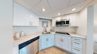 Located at 2500 Edwards Dr in Fort Myers, Florida, this coastal-inspired space offers a thoughtfully designed single-floor layout with a relaxed, beach-themed aesthetic throughout. The living area centers around a large tufted navy sectional sofa accented with coral and starfish throw pillows, complemented by a pink accent chair and light wood flooring. Nautical wall art featuring seashell and ocean motifs reinforces the seaside ambiance. The kitchen is well-equipped, featuring warm medium-tone wood cabinetry, granite-style countertops, a stainless steel sink with a pull-down faucet, and a stainless steel dishwasher, providing ample storage and functionality. The primary bedroom is a standout, offering floor-to-ceiling windows that frame breathtaking panoramic waterfront views, filling the room with natural light. It is furnished with a rattan-style bed dressed in a vibrant coastal-patterned quilt. A second bedroom features a similar coastal décor with warm, carpeted flooring and a cozy, well-appointed layout. The bathroom is finished with warm wood vanity cabinetry, light stone-toned countertops, tile flooring, and a glass-enclosed walk-in shower with subway tile surround. Together, these spaces create an inviting, resort-like atmosphere ideal for enjoying the stunning Florida waterfront setting.