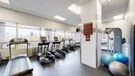 Located at 195 Clearview Ave in Ottawa, Ontario, this well-equipped fitness room offers a comprehensive workout facility within a efficiently organized single-level space. The room features a drop-tile ceiling with recessed panel lighting, durable rubber-composite flooring, and a continuous band of large windows along one wall that floods the space with natural light and provides views of the surrounding outdoor greenery.

The cardio section is well-stocked with multiple treadmills, elliptical trainers, and stationary bikes, all positioned to take advantage of the natural light and outdoor views. A dedicated strength-training area includes a full dumbbell rack, a flat workout bench, a cable resistance machine, and wall-mounted resistance equipment. Stability balls are also available for functional training exercises.

A large wall-mounted mirror on one end visually expands the space and allows users to monitor their form during workouts, while a flat-screen television provides entertainment during sessions. The room is accessed through a glass-paneled sliding door, and motivational athletic artwork adorns the walls, contributing to an energizing atmosphere. A water fountain is conveniently accessible within the space. Posted fitness room rules ensure a safe and respectful environment for all users.