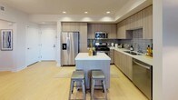 This single-floor residence showcases a thoughtfully designed layout with contemporary finishes throughout. The kitchen is a standout feature, equipped with stainless steel appliances including a gas range, built-in microwave, and French door refrigerator. Flat-front cabinetry in a warm wood-grain finish extends to the ceiling, complemented by a gray subway tile backsplash and clean white quartz countertops. A central island with seating provides additional prep space and casual dining functionality. Light hardwood-style flooring runs through the main living areas, enhancing the open feel of the space.

The living area is bright and inviting, with large windows and a sliding glass door opening to an outdoor balcony, offering views of the surrounding neighborhood. Natural light fills the room, highlighting the light-toned flooring and neutral wall palette.

The bedrooms feature plush carpeting, ceiling fans, and windows that allow for natural ventilation and light. The bathroom includes a full tub-shower combination with tile surround, open shelving, a vanity, and light wood-style flooring that ties the space together cohesively. The overall aesthetic is clean, modern, and functional throughout.
