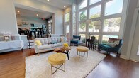 This thoughtfully designed single-floor space presents a cohesive blend of modern aesthetics and functional layout, highlighted by a warm teal, gray, and gold color palette carried consistently throughout each area. The main lounge is a standout feature, boasting soaring double-height ceilings lined with expansive floor-to-ceiling windows that flood the interior with natural light and offer tree-lined outdoor views. Rich hardwood flooring anchors the space, complementing the plush white area rug, tufted gray sofa, and gold velvet ottomans arranged around a live-edge coffee table.

A dramatic teal accent wall frames a stacked-stone fireplace topped with a wall-mounted television, creating a striking focal point in the living area. Adjacent to the main lounge, a sun-drenched dining area features high-top tables with industrial metal seating, positioned beside a wall of French doors and transom windows that open to the outdoors.

The kitchenette is equipped with stainless steel appliances, granite countertops, a herringbone tile backsplash, open iron shelving, and a utility sink — designed for practical use. A dedicated alcove workspace with a built-in desk and teal accent wall completes the functional offerings of this well-appointed space.