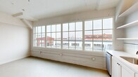 Located at 2154 Noisette Blvd in North Charleston, SC, this one-bedroom, one-bathroom unit showcases an industrial loft aesthetic rooted in the building's architectural character. The interior is finished throughout in crisp white tones, creating a clean, open atmosphere that maximizes the sense of space.

The bedroom is generously proportioned, featuring exposed ceiling ductwork and piping that highlight the unit's loft-style design, along with built-in closets with classic panel doors. Large multi-pane factory-style windows flood the room with natural light and offer views of the surrounding North Charleston streetscape, including the area's historic brick buildings.

The kitchen is efficiently laid out along a single wall and includes white shaker-style cabinetry, open floating shelves, a subway tile backsplash, and a full suite of stainless steel appliances — including a refrigerator, range, microwave, and dishwasher. A window adjacent to the kitchen further brightens the space.

The hallway connects the main living areas to the bathroom, which features classic white tile flooring with a dark accent border, a glass-enclosed walk-in shower with subway tile, matte black fixtures, and a white vanity. The overall ambiance of the unit is bright, modern, and industrial-inspired.