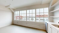 Located at 2154 Noisette Blvd in North Charleston, SC, this one-bedroom, one-bathroom unit showcases an industrial loft aesthetic rooted in the building's architectural character. The interior is finished throughout in crisp white tones, creating a clean, open atmosphere that maximizes the sense of space.

The bedroom is generously proportioned, featuring exposed ceiling ductwork and piping that highlight the unit's loft-style design, along with built-in closets with classic panel doors. Large multi-pane factory-style windows flood the room with natural light and offer views of the surrounding North Charleston streetscape, including the area's historic brick buildings.

The kitchen is efficiently laid out along a single wall and includes white shaker-style cabinetry, open floating shelves, a subway tile backsplash, and a full suite of stainless steel appliances — including a refrigerator, range, microwave, and dishwasher. A window adjacent to the kitchen further brightens the space.

The hallway connects the main living areas to the bathroom, which features classic white tile flooring with a dark accent border, a glass-enclosed walk-in shower with subway tile, matte black fixtures, and a white vanity. The overall ambiance of the unit is bright, modern, and industrial-inspired.