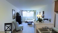 This single-floor unit is arranged in a long, rectangular layout that organizes living, dining, kitchen, and sleeping areas in a continuous flow. The living room serves as a central gathering space, finished with plush carpeting and crisp white walls that create a bright, airy atmosphere. A large window dressed with decorative valance curtains allows generous natural light to fill the room, enhancing the clean, neutral palette. Seating is arranged around a round coffee table, with additional accent chairs and storage furniture complementing the space.

The kitchen is positioned toward one end of the unit, featuring warm wood cabinetry and a countertop that partially opens toward the living area, providing a convenient pass-through feel. A sink and standard appliances complete the functional cooking space.

Toward the opposite end, a sleeping area is visible in the floor plan, separated from the main living zone to provide privacy. Throughout the unit, the consistent use of light-toned walls and carpeted flooring unifies the overall aesthetic, creating a cohesive and comfortable environment. The layout makes efficient use of the available space while maintaining a sense of openness and practicality.