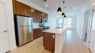 This well-appointed single-floor residence showcases a contemporary design with a cohesive blend of warm and neutral tones throughout. The heart of the space is an open-concept kitchen, where rich walnut-toned cabinetry extends floor to ceiling, providing generous storage alongside a full suite of stainless steel appliances, including a refrigerator, electric range, and built-in microwave. A large central island with a crisp white quartz countertop and an integrated sink serves as both a functional workspace and a natural gathering point. Stylish matte black pendant lights with warm gold interiors hang above, adding a modern, sophisticated touch. Light hardwood flooring flows seamlessly through the kitchen and living areas, contributing to a bright, airy ambiance enhanced by large windows that invite abundant natural light. The bedroom is a calm and neutral retreat, finished with plush carpeting, white walls, and window blinds for privacy and light control. The bathroom continues the space's design language with wood-toned cabinetry, white quartz countertops, light hardwood-style flooring, and a full tub-shower combination, complemented by a walk-in closet accessible directly from the bathroom. The overall aesthetic is clean, modern, and move-in ready.
