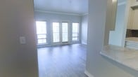 This single-floor space offers a clean, modern aesthetic defined by an open living area with light gray hardwood-style flooring that runs seamlessly throughout. The room is anchored by a set of French doors flanked by large windows, allowing generous amounts of natural light to fill the interior and creating an airy, welcoming atmosphere. The soft, neutral wall tones complement the flooring and contribute to the space's understated elegance.

Crown molding adds a refined architectural detail along the ceiling line, elevating the overall finish of the room. White baseboard trim provides a crisp contrast against the pale walls, reinforcing the clean, contemporary feel of the space. A partial wall or countertop edge visible at the right side of the frame suggests an adjacent area, potentially a kitchen or bar counter, hinting at a practical open-concept layout.

The living area is generously proportioned, offering flexible arrangement possibilities. The abundance of natural light, neutral color palette, and quality finishes combine to create a bright, comfortable environment that feels both polished and inviting throughout the day.