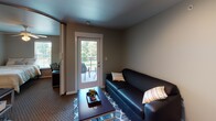 Located at 1201 Laporte Ave in Valparaiso, Indiana, this single-floor unit offers a well-organized layout that efficiently combines living, dining, sleeping, and kitchen functions. The living area features neutral-toned walls, carpeted flooring, and a glass-paned door that opens to a private outdoor balcony, allowing natural light to filter in and providing views of surrounding greenery. A dark leather sofa and coffee table anchor the space comfortably.

Adjacent to the living area is a compact dining nook accommodating a round dark-wood table with seating for several, suitable for casual meals. The kitchen is outfitted with white cabinetry, granite-style countertops, and black appliances including a refrigerator, electric range, over-the-range microwave, and a stainless steel sink — providing all essential cooking amenities in a practical arrangement. Wood-style flooring distinguishes the kitchen from the carpeted living spaces.

The bedroom is a comfortable retreat featuring a ceiling fan with lighting, a window with outdoor views, and dedicated study space with a desk and shelving unit. The bathroom is finished with tile flooring, a granite-top vanity with dark cabinetry, a glass-enclosed shower stall, and clean white walls, offering a functional and tidy layout throughout.