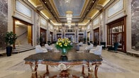 Located at 2300 N Lincoln Park West in Chicago's Lincoln Park neighborhood, this magnificent lobby is a showcase of beaux-arts grandeur. The expansive, double-height space commands attention with towering columns clad in richly veined purple and gray marble, set against cream-toned stone walls accented with intricate gold detailing. A stunning tiered crystal chandelier anchors the ornately decorated ceiling, which features painted murals framed by gilded molding, casting a warm and luminous glow throughout the hall. Wrought-iron balconies with decorative railings overlook the main floor, adding a sense of vertical drama to the already soaring interior. The floor is laid in elegantly patterned light stone tile, complementing the refined material palette. Seating areas are thoughtfully arranged throughout, featuring contemporary gray sofas, tufted lounge chairs, and plush emerald velvet wingback chairs that offer an inviting contrast to the classical architecture. A stately ornamental console table with gilded carvings serves as a focal centerpiece. Tall mahogany-framed glass doors and windows connect the various areas, allowing light to flow freely. A marble reception desk and a graceful staircase with brass railings complete this distinguished and timeless space.