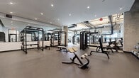 This expansive fitness center is thoughtfully designed to accommodate a wide range of training disciplines, blending industrial aesthetics with a clean, modern atmosphere. The space is divided into distinct functional zones, each outfitted with professional-grade equipment to support varied workout styles.

The strength training area features a polished concrete floor, floor-to-ceiling mirrors that amplify the sense of space, and recessed lighting overhead. It is stocked with power racks, cable machines, weight benches, and a comprehensive selection of free weights, including kettlebells and medicine balls organized on dedicated racks.

Transitioning into the cardio zone, the design shifts to an industrial character with exposed concrete ceilings, bold black ductwork, and structural columns. Generous floor-to-ceiling windows flood this area with natural light and offer views to the exterior. Equipment here includes stair climbers, ellipticals, treadmills, rowing machines, and stationary bikes from brands such as StairMaster and Freemotion.

A dedicated functional training area features a RaX multi-use rig with suspension trainers, battle ropes, and a variety of accessories, set on a padded rubberized floor with marked training zones. The facility connects seamlessly between zones through steel-framed glass doors, maintaining visual openness throughout. The overall ambiance is energetic yet refined, making it a well-rounded destination for serious fitness.