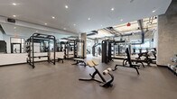 This expansive fitness center is thoughtfully designed to accommodate a wide range of training disciplines, blending industrial aesthetics with a clean, modern atmosphere. The space is divided into distinct functional zones, each outfitted with professional-grade equipment to support varied workout styles.

The strength training area features a polished concrete floor, floor-to-ceiling mirrors that amplify the sense of space, and recessed lighting overhead. It is stocked with power racks, cable machines, weight benches, and a comprehensive selection of free weights, including kettlebells and medicine balls organized on dedicated racks.

Transitioning into the cardio zone, the design shifts to an industrial character with exposed concrete ceilings, bold black ductwork, and structural columns. Generous floor-to-ceiling windows flood this area with natural light and offer views to the exterior. Equipment here includes stair climbers, ellipticals, treadmills, rowing machines, and stationary bikes from brands such as StairMaster and Freemotion.

A dedicated functional training area features a RaX multi-use rig with suspension trainers, battle ropes, and a variety of accessories, set on a padded rubberized floor with marked training zones. The facility connects seamlessly between zones through steel-framed glass doors, maintaining visual openness throughout. The overall ambiance is energetic yet refined, making it a well-rounded destination for serious fitness.