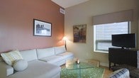 This single-floor space offers a thoughtfully designed interior that balances modern aesthetics with warm, inviting elements. The living area serves as the focal point of the space, showcasing notably high ceilings that amplify the sense of openness and volume. A rich terracotta-toned accent wall adds depth and warmth, complementing the neutral cream and beige tones found throughout the rest of the room. Natural light filters in through a large window fitted with classic horizontal blinds, brightening the space and enhancing its airy feel. The living area is furnished with a generous white sectional sofa, anchored by a patterned green area rug and a sleek glass coffee table that adds a contemporary touch. Warm ambient lighting from a classic floor lamp contributes to the cozy atmosphere during evening hours. The walls are adorned with framed artwork, adding a curated, gallery-like quality to the space. Light hardwood flooring runs throughout, tying together the design elements cohesively. Woven storage baskets provide both functional and decorative accents. Overall, the space presents a clean, modern interior with a warm residential character, ideal for comfortable everyday living and entertaining.