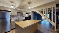 Located at 1503 Monmouth Avenue in Lancaster, Ohio, this two-floor home offers a thoughtfully updated interior with a cohesive modern design throughout. The kitchen serves as a standout feature, showcasing gray shaker-style cabinetry, granite countertops, and a mosaic tile backsplash in cool blue and gray tones. A large butcher block island anchors the space, providing ample prep and gathering room, while pendant lighting suspended from exposed beam detailing above adds an industrial-chic character. Stainless steel appliances, including a refrigerator, range, and dishwasher, complete the functional layout. Light wood-tone flooring runs through the main living areas, complementing the neutral gray palette used throughout the home. A staircase connects the two levels, with sliding glass door access visible from the kitchen area, bringing in natural light. The bathrooms feature gray cabinetry with granite-style countertops, light wood flooring, and a tub-shower combination. The bedrooms are carpeted in a soft gray tone and equipped with ceiling fans and natural light from windows overlooking greenery outside. The textured ceilings and consistent gray wall tones tie the entire home together with a clean, contemporary aesthetic.