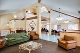Located at 12801 Rosecrans Ave in Norwalk, California, this single-story community clubhouse offers a welcoming and versatile gathering space designed for social connection and shared activities. The interior is finished in warm yellow tones throughout, complemented by vaulted ceilings with exposed wood beams that lend architectural character and a sense of openness to the space.

The main lounge area serves as the heart of the facility, featuring a classic white-mantled fireplace with dark marble surround, plush leather seating, and a green sofa arrangement centered around a glass-top coffee table. This area flows seamlessly into an entertainment nook equipped with a large flat-screen television, audio components, and additional leather seating — all bathed in natural light from large grid-paned windows that frame views of the surrounding landscape.

Adjacent to the lounge, a spacious dining area accommodates multiple round tables with black chairs beneath an elegant chandelier and vaulted ceiling, making it well-suited for community meals and events. The warm hardwood flooring in this section contrasts with the teal tile entryway, adding visual interest to the open layout.

A dedicated music and activity room rounds out the space, complete with an upright piano, a writing desk, and ample carpet area, all surrounded by large windows that invite natural light. The cohesive design and multi-functional layout make this clubhouse an inviting hub for community life.