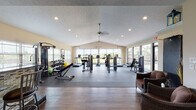 Located at 1000 Cassandra Ln in La Grange, Kentucky, this single-story fitness center offers a well-appointed and functional facility designed to support a comprehensive workout experience. The main exercise area is expansive, featuring vaulted ceilings with dual ceiling fans, recessed lighting, and a textured ceiling finish. Large windows along multiple walls flood the space with natural light while providing open views of the surrounding landscape, creating an energizing atmosphere. The rubber-floored workout zone is equipped with a wide array of exercise machines, including treadmills, stationary bikes, ellipticals, cable machines, and free weights, accommodating a full range of fitness needs. A welcoming entry lounge transitions from the main entrance into the workout area, featuring hardwood-style flooring, wicker seating, and interior doors leading to additional rooms. The kitchenette area is a standout feature, showcasing rich wood cabinetry, dark granite countertops, a mosaic tile backsplash, and a raised bar with barstool seating — ideal for post-workout refreshment and socializing. Black appliances including a refrigerator and microwave complete the functional kitchen setup. Throughout the facility, warm neutral wall tones and thoughtful design details contribute to a clean, welcoming, and professional environment.