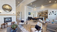 This elegantly designed single-floor space offers an open-concept layout that seamlessly connects multiple functional areas under soaring ceilings. The centerpiece of the main living area is a floor-to-ceiling whitewashed stone fireplace with a rustic wood mantel and a decorative round mirror above, creating a striking focal point. Stone-tiled and light wood flooring flows throughout, grounding the space with a warm, contemporary aesthetic.

The lounge area features multiple seating arrangements bathed in natural light from large, arched windows and full-length paned glass that frame outdoor greenery views. A dedicated dining nook, enclosed by dramatic arched windows with elegant navy and white drapery, provides an intimate setting with abundant daylight.

The kitchen area is outfitted with a thick marble-topped island, dark stainless steel appliances, open natural wood floating shelves, hexagonal tile backsplash, and matte black pendant lighting. A separate seating zone with its own dining table and media display offers additional gathering space.

Throughout, textured accent walls, recessed lighting, and crown molding add refined architectural detail. The overall ambiance is modern yet inviting, blending natural materials with contemporary finishes to create a polished, versatile communal environment.