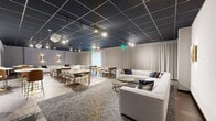 This contemporary multipurpose space offers a well-appointed, single-floor layout designed for flexible use across meetings, gatherings, and events. The room is defined by a striking dark grid-panel ceiling fitted with adjustable track lighting, which provides targeted illumination throughout and creates a sleek, modern ambiance. Floor-to-ceiling drape panels line the walls, lending a soft, refined aesthetic while adding acoustic warmth to the space.

The layout is thoughtfully divided into two primary functional zones. A comfortable lounge area features large, neutral-toned sectional sofas arranged around paired gold-accented coffee tables atop a textured area rug, ideal for informal gatherings or breakout conversations. Adjacent to this, a dining and meeting area provides multiple rectangular wooden tables with upholstered chairs, accommodating groups of varying sizes. Smaller round tables with bar-height seating offer additional flexibility.

A retractable projection screen is mounted on one wall, paired with a ceiling-mounted projector, making the space well-suited for presentations or screenings. Elegant brass wall sconces add a warm decorative touch, while light wood-look flooring anchors the dining zone and transitions seamlessly into the carpeted lounge. Additional doors suggest access to adjoining rooms or facilities, further enhancing the space's utility for a wide range of events.