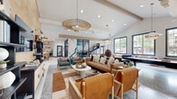 This expansive single-floor amenity space is thoughtfully designed to encourage gathering and recreation, combining bold aesthetics with a warm, inviting atmosphere. Soaring vaulted ceilings with exposed ductwork and wooden beams lend an industrial-chic character, while large dark-framed windows flood the interior with abundant natural light and views of surrounding greenery.

The open-plan layout flows seamlessly between several distinct zones. A comfortable lounge area anchors the space, furnished with a mix of leather and plaid upholstered seating, layered rugs, and sculptural side tables, all centered beneath striking oversized rattan pendant lights. A textured geometric feature wall in warm wood tones serves as a dramatic backdrop for a mounted flat-screen TV and flanking open shelving.

Adjacent to the lounge, a game area offers a foosball table and a skee-ball machine, set against the backdrop of a bold painted mural and large windows overlooking the outdoors. A glass-partitioned private room provides a more intimate setting, visible through striped frosted glass doors. A kitchen and dining area with open shelving completes the space.

Terrazzo flooring throughout ties the areas together, adding a sophisticated yet playful design element. The overall ambiance is modern, energetic, and community-focused.
