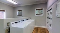This single-floor shared laundry facility is designed for practicality and comfort. The room is finished with warm hardwood flooring that adds a welcoming touch to an otherwise utilitarian space. A row of commercial-grade top-loading washing machines lines one wall, providing ample capacity for high-volume laundry needs. The walls are painted in a neutral gray tone, lending the space a clean and modern appearance, while crisp white baseboards and crown molding add a polished architectural detail. Two windows dressed with natural bamboo roller shades allow soft, diffused natural light to brighten the room, reducing reliance on the overhead fluorescent lighting. Ventilation is supported by ceiling vents, ensuring the space remains comfortable during use. The open layout provides easy access to all machines and allows for unobstructed movement throughout the room. Overall, the facility balances functional design with thoughtful finishing touches, making it a well-maintained and inviting space for everyday laundry use.