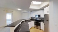 This single-floor space showcases a thoughtfully designed kitchen that serves as its central feature. The kitchen is outfitted with a full suite of stainless steel appliances, including an electric range with oven, an over-the-range microwave, and a refrigerator, all contributing to a sleek, modern look. White shaker-style cabinetry lines the walls, providing ample storage, while dark charcoal countertops create a striking contrast against the bright cabinetry and light-toned hardwood flooring. An L-shaped counter configuration, complete with an undermount sink, maximizes workspace efficiency. A peninsula-style counter extension adds additional prep and seating space, enhancing the kitchen's functionality. The ceiling features a raised, stepped detail with a centrally positioned flush-mount light fixture, adding architectural dimension to the space. Recessed lighting and additional ceiling-mounted fixtures ensure the area is well-lit throughout. The open layout connects the kitchen to an adjacent living area, creating a cohesive, airy flow across the unit. The overall aesthetic is clean, contemporary, and functional.