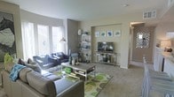 This single-story space centers around a well-appointed living area designed for both comfort and functionality. The room is finished with neutral-toned carpet and painted in soft, warm gray tones that create a calming backdrop throughout. Natural light enters generously through a large window along one wall, brightening the space and highlighting the contemporary furnishings.

The living area is anchored by a plush gray sectional sofa and a coordinating armchair, arranged around a sleek chrome and wood coffee table atop a green-patterned area rug. A chrome-framed open shelving unit displays decorative accents in shades of blue and white, adding visual interest and personality to the room.

A built-in media niche on the opposite wall provides a dedicated space for a flat-screen television, flanked by a modern console table and decorative elements. Above, a grouping of framed botanical artwork in blue and green tones ties the color palette together cohesively.

A hallway visible toward the rear of the space suggests additional rooms beyond, contributing to the overall sense of a functional, connected floor plan. The space presents a clean, modern aesthetic with thoughtful styling throughout.