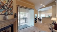 This single-floor residence offers a thoughtfully designed open-concept layout that connects the living, dining, and kitchen areas in a cohesive and functional flow. The living area is anchored by a classic fireplace, adding warmth and character to the space, while plush carpeting underfoot contributes to a comfortable, inviting atmosphere. Warm neutral tones on the walls complement the overall design, creating a welcoming ambiance throughout.

Large sliding glass doors fitted with vertical blinds open directly onto a private balcony, flooding the interior with natural light and providing an easy indoor-outdoor connection. The balcony overlooks a lush, tree-lined outdoor area, offering a serene retreat.

Adjacent to the living space, the dining area is well-defined yet open, featuring a dining set and a nearby bookcase for added functionality. Track lighting runs along the ceiling in this area, providing versatile and practical illumination. The kitchen is visible beyond, marked by a raised countertop bar that serves as a natural dividing element while maintaining the open feel of the space. A window in the dining and kitchen area further enhances natural light. Together, these elements create a balanced and comfortable single-floor home.