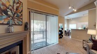 This single-floor residence offers a thoughtfully designed open-concept layout that connects the living, dining, and kitchen areas in a cohesive and functional flow. The living area is anchored by a classic fireplace, adding warmth and character to the space, while plush carpeting underfoot contributes to a comfortable, inviting atmosphere. Warm neutral tones on the walls complement the overall design, creating a welcoming ambiance throughout.

Large sliding glass doors fitted with vertical blinds open directly onto a private balcony, flooding the interior with natural light and providing an easy indoor-outdoor connection. The balcony overlooks a lush, tree-lined outdoor area, offering a serene retreat.

Adjacent to the living space, the dining area is well-defined yet open, featuring a dining set and a nearby bookcase for added functionality. Track lighting runs along the ceiling in this area, providing versatile and practical illumination. The kitchen is visible beyond, marked by a raised countertop bar that serves as a natural dividing element while maintaining the open feel of the space. A window in the dining and kitchen area further enhances natural light. Together, these elements create a balanced and comfortable single-floor home.
