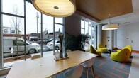 This contemporary single-floor space combines thoughtful design with a welcoming, functional atmosphere. Large floor-to-ceiling windows line the perimeter, flooding the interior with natural light and offering open views of the surrounding streetscape. The warm wood-paneled ceiling adds a natural contrast to the modern architectural elements, creating a balanced and inviting ambiance throughout.

The main area features a generous communal table with clean-lined white chairs, ideal for meetings or collaborative work. Overhead, a distinctive dome-shaped pendant light anchors the space, complementing additional modern lighting fixtures nearby. Lush indoor plants are strategically placed along the window line, introducing a refreshing natural element that softens the contemporary aesthetic.

A lounge area set slightly apart offers a change of pace, furnished with sculptural, chartreuse accent chairs that provide a pop of color against the patterned carpet flooring. The combination of a central work table and relaxed seating zones makes this space adaptable for a variety of uses, from focused work sessions to informal gatherings. The overall design reflects a modern, professional sensibility with carefully selected furnishings and finishes that prioritize both style and functionality.
