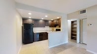 This single-floor space showcases a well-appointed kitchen as its centerpiece, designed with both functionality and style in mind. The kitchen features rich, dark espresso-toned cabinetry that extends along the back wall, providing ample upper and lower storage. Black appliances, including a full-size refrigerator, dishwasher, and range, complement the dark cabinetry for a cohesive aesthetic. A light-toned countertop offers a striking contrast, and a double sink is positioned beneath the upper cabinets. Recessed lighting illuminates the kitchen area effectively, highlighting the clean lines of the design. A partial knee wall creates a subtle division between the kitchen and the adjacent open area, adding architectural interest while maintaining an open, connected feel. Light-toned ceramic tile flooring runs throughout the visible space, contributing to a bright and airy atmosphere. To the right of the kitchen, a built-in open shelving unit provides additional storage or display space, seamlessly integrated into the layout. The neutral wall tones and clean finishes throughout give the space a modern, move-in-ready appeal.