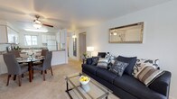 This single-floor residence showcases an open-concept design that seamlessly integrates the living room, dining area, and kitchen into one cohesive space. The living room serves as the central hub, featuring plush carpeted flooring, a ceiling fan for year-round comfort, and ample room for comfortable furnishings. A glass-top coffee table and a deep navy sofa with decorative pillows contribute to the space's modern, understated aesthetic. A framed mirror on the wall enhances the sense of openness by reflecting light throughout the room.

The dining area is positioned adjacent to the living space, providing a natural transition and accommodating a small table with seating. The kitchen is efficiently designed with white cabinetry, a full-sized refrigerator, and a breakfast bar that doubles as additional counter space and a casual dining option. The white color palette of the kitchen keeps the area feeling bright and clean.

A hallway visible from the living room leads to additional areas of the unit, suggesting a practical and well-organized floor plan. Overall, the space offers a comfortable, functional layout suited for everyday living.