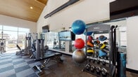This fitness center is a thoughtfully designed, single-floor facility that combines modern architecture with a full suite of workout equipment. The space is defined by its striking vaulted ceiling, which features warm wood paneling and exposed industrial-style ductwork, lending the room an open, airy character. Large floor-to-ceiling windows along the perimeter flood the interior with natural light and provide views of the surrounding exterior, creating an energizing atmosphere for exercise.

The flooring throughout consists of a patterned tile-style carpet in a checkerboard design, adding visual interest while providing a durable, functional surface. A ceiling fan with recessed lighting ensures comfortable airflow across the main workout area.

The facility is comprehensively equipped for a wide range of training styles. Cardio equipment, including treadmills, ellipticals, and stationary bikes, lines one side of the room. The strength training section features cable machines, a multi-station weight system, and adjustable benches. A dedicated functional training area includes TAG-branded ball racks stocked with medicine balls and stability balls, along with a full rack of dumbbells and kettlebells in varying weights.

Floor-to-ceiling mirrors expand the sense of space and allow users to monitor their form. A built-in white cabinetry unit with a countertop provides storage for towels and accessories, rounding out this well-appointed fitness space.