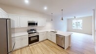 This single-floor residence offers a thoughtfully designed layout comprising one bedroom, one bathroom, a kitchen, and a living room. The open-concept kitchen is a standout feature, showcasing white shaker-style cabinetry, crisp white countertops, and a full suite of stainless steel appliances including a refrigerator, electric range, over-the-range microwave, and dishwasher. A peninsula with an undermount sink and stylish brushed nickel pendant lights above anchors the space, making it ideal for both cooking and casual dining. The kitchen flows seamlessly into the living area, which is finished with warm-toned hardwood-style flooring that extends throughout the main living spaces, creating a cohesive and inviting atmosphere. The bedroom is carpeted for comfort and features a ceiling fan with a light fixture, a window with a roller shade that admits natural light and an exterior view, and a spacious walk-in closet with built-in shelving for organized storage. The bathroom is finished with light hardwood-style flooring, a white vanity with ample cabinet storage, a large mirror, and a combined tub and shower. Neutral warm-beige walls and clean white trim carry consistently throughout the residence, contributing to a bright, modern, and move-in-ready aesthetic.