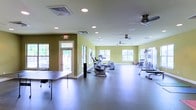 This single-floor fitness center offers a spacious, open layout designed to accommodate a variety of workout activities. The room features light green walls that contribute to an energizing atmosphere, complemented by large windows along multiple walls that allow abundant natural light to filter in, creating a bright and welcoming environment. The polished dark floor provides a durable, easy-to-maintain surface suitable for both equipment use and open exercise areas. Overhead, multiple ceiling fans work alongside recessed lighting to ensure consistent air circulation and illumination throughout the space. The fitness area is equipped with a selection of cardio machines, including stationary bikes, an elliptical, and a stair climber, as well as a cable strength training unit positioned toward the far end of the room. A folding table near the entrance adds functional versatility to the space. A clearly marked exit door with exterior window access provides both safety and additional natural light. The generous open floor area allows for flexible exercise arrangements, making the space suitable for group activities or individual workout routines.