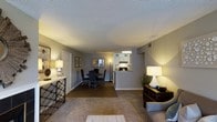 Located at 2600 Waterford Pl in Hoover, Alabama, this single-floor residence offers a thoughtfully designed open-concept layout that seamlessly connects the living, dining, and kitchen areas. The living room is generously proportioned, featuring warm carpeted flooring, neutral gray walls, and a textured ceiling that adds dimension overhead. Ambient lighting from stylish table lamps creates a welcoming atmosphere, complemented by a classic console table that anchors the entryway. The dining area sits centrally within the open floor plan, accommodating a full dining set beneath soft overhead lighting, making it ideal for both everyday meals and entertaining. A pass-through window connects the dining space to the kitchen, which features white cabinetry, under-cabinet lighting, and a functional layout that maximizes efficiency. The kitchen's bright, clean aesthetic contrasts pleasantly with the warmer tones of the living and dining areas. Throughout the space, the cohesive color palette of soft grays and warm neutrals lends a calm, unified ambiance. The single-floor design ensures easy accessibility and a smooth flow between all areas, making this Hoover residence a practical and comfortable living environment.