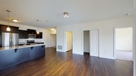 This single-floor space showcases an open-concept layout that integrates the kitchen and living area into one cohesive environment. Rich, dark hardwood flooring runs throughout, providing a warm and grounding aesthetic that contrasts cleanly with the bright white walls and ceilings.

The kitchen is outfitted with dark espresso cabinetry, stainless steel appliances including a range and microwave, and a peninsula with seating overhang — ideal for casual dining or entertaining. Pendant lighting above the peninsula adds a modern accent, complementing the recessed-style ceiling fixtures in the living area.

The open living space is generously sized, allowing for flexible furniture arrangements. Two interior doors are visible — one leading to what appears to be a hallway or additional room, and another that may serve as a closet, adding functional storage to the layout.

Natural light enters through the space, and the clean, neutral palette throughout ensures a bright and airy atmosphere. The overall design is contemporary and practical, with finishes that balance style and durability seamlessly.
