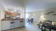 This single-floor residence showcases an open-concept design that seamlessly integrates the kitchen, dining, and living areas into one cohesive, flowing space. The kitchen is equipped with warm wood-toned cabinetry, a stainless steel range with overhead ventilation, and a raised breakfast bar with bar stool seating — ideal for casual dining or entertaining. Pendant lighting above the bar adds a focused, stylish touch to the workspace.

Adjacent to the kitchen, the dining area accommodates a full dining table and chairs, illuminated by a decorative chandelier that provides warm ambient light. A decorative sunburst mirror accents the neutral-toned wall, adding visual interest to the space. Natural light flows in through a nearby window, contributing to the bright and airy atmosphere throughout.

The living area extends naturally from the dining space, featuring soft carpeting underfoot and neutral wall tones that create a calm, welcoming environment. A floor lamp provides supplemental lighting, and the layout allows for flexible furniture arrangements. Overall, the space offers a practical and comfortable open-plan living experience.