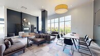 Located at 111 Towne St in Stamford, CT, this single-floor amenity space offers a generously sized, open-concept layout designed for both comfort and social engagement. The living area is defined by soaring ceilings, large floor-to-ceiling windows that flood the space with natural light, and sweeping outdoor views of lush greenery. Multiple seating arrangements — including modular sofas, lounge chairs, and café-style tables — create distinct zones for relaxing or gathering. A sleek linear fireplace anchors one wall, accented by decorative round mirrors and artistic sculptural elements, lending a refined, contemporary ambiance. A large flat-screen television and ample flexible seating make the lounge equally suited for casual entertainment. The kitchen features high ceilings, bold cobalt-blue walls, white shaker cabinetry, granite countertops, a substantial center island with bar seating, stainless steel appliances, and direct access to an outdoor terrace. Throughout the space, a vivid geometric-patterned carpet adds a dynamic, modern aesthetic, while curated artwork lines the hallways. A self-service market area with snack and beverage stations provides added convenience. Recessed lighting and oversized pendant fixtures complete the polished, welcoming atmosphere of this thoughtfully designed communal space.