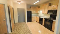 This single-floor residential unit offers a practical and well-organized layout designed for comfortable everyday living. The kitchen is a standout feature, equipped with light natural wood cabinetry that lines the walls in an efficient L-shaped configuration, providing ample storage space. Black appliances — including a full-size refrigerator, gas range, and dishwasher — offer a clean contrast against the light wood tones and neutral countertops. A double basin sink is positioned beneath upper cabinetry, maximizing workspace and utility. Overhead fluorescent lighting ensures the kitchen is well-lit for meal preparation. The flooring transitions from smooth light wood laminate in the kitchen area to a checkered tile pattern near the entryway, adding visual distinction between zones. The entry corridor features a solid, gray steel door, lending a secure and sturdy feel to the unit's access point. A flush-mount ceiling light fixture in the entryway provides functional illumination. The overall ambiance of the space is clean, straightforward, and move-in ready, with neutral wall tones and durable finishes throughout that are well-suited for practical, day-to-day living.