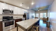 This well-appointed 3-bedroom, 3-bathroom single-floor residence offers a thoughtfully designed layout that maximizes both function and style. The open-concept kitchen and living area serves as the heart of the home, featuring hardwood flooring, a striking exposed corrugated metal ceiling, and large windows that frame urban views and invite abundant natural light. The kitchen is equipped with stainless steel appliances, white upper cabinetry paired with dark lower cabinets, granite countertops, and a large central island with bar seating — ideal for both meal prep and casual dining. The adjoining living area provides a comfortable, open atmosphere beneath the distinctive industrial ceiling detail. Each of the three bedrooms features carpeted flooring and windows overlooking the surrounding cityscape, offering a quiet retreat. The primary bedroom includes access to a closet. The three bathrooms are consistently finished with tile flooring, granite-topped dark wood vanities, and full tub-shower combinations. Two dedicated closets provide additional storage throughout the unit. The overall design blends modern finishes with industrial architectural elements, creating a cohesive and contemporary urban living environment.