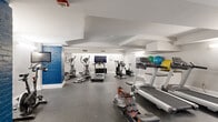 Located at 1211 West 9th Street in Cleveland's vibrant Ohio City neighborhood, this fitness center offers a thoughtfully designed exercise space across a single floor. The room is finished with crisp white walls and a durable rubber-speckle floor, creating a clean, functional environment suited for a range of workout routines. A standout architectural detail is the bold blue painted brick accent column, which adds a distinctive industrial character to the otherwise modern interior. The ceiling features exposed structural beams painted white, contributing to the room's defined layout and zoned feel. The space is equipped with multiple treadmills, elliptical trainers, a stationary spin bike, an air resistance bike, a rowing machine, and a wall-mounted flat-screen TV to support entertainment during cardio sessions. A dedicated stretching and cool-down area includes an exercise mat and a wall-mounted shelf displaying stability balls and medicine balls in various sizes. Accent lighting along the walls provides even illumination throughout, while a large interior window connects to an adjacent area, allowing natural light to filter in. The overall ambiance is energetic yet organized, offering residents a comprehensive fitness facility without leaving the building.